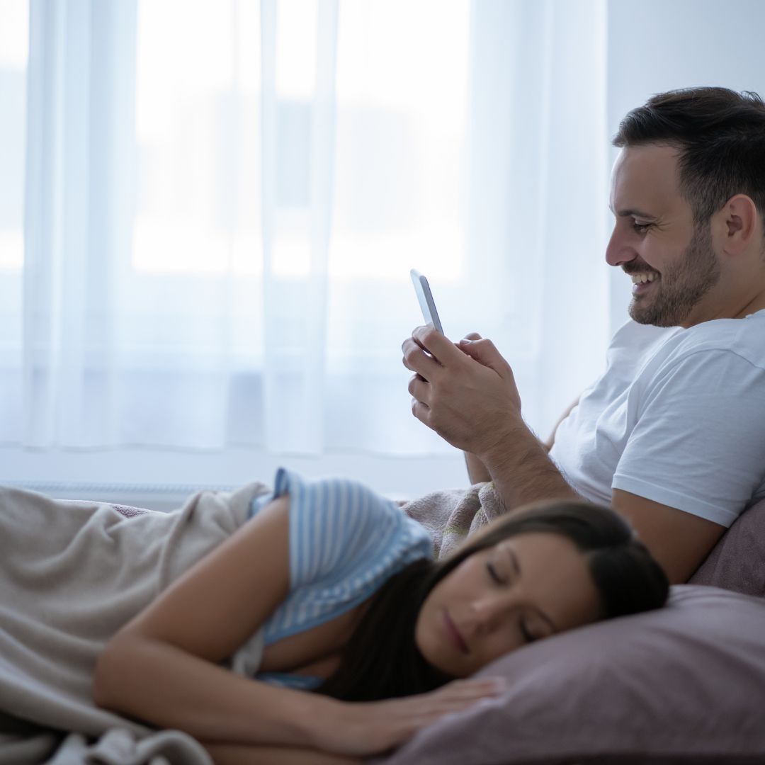 Zdjęcie przedstawia mężczyznę uśmiechającego się do telefonu podczas gdy jego partnerka śpi obok, co może sugerować tematykę zdrady i radykalnego wybaczenia.